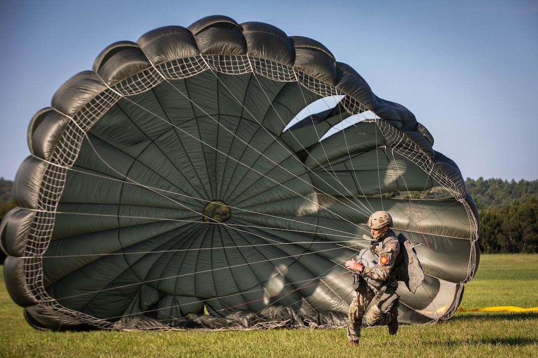A soldier wearing a parachute runs in a field during the day.