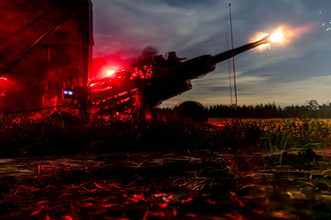 Soldiers fire a howitzer in a field in the dark illuminated by red light.