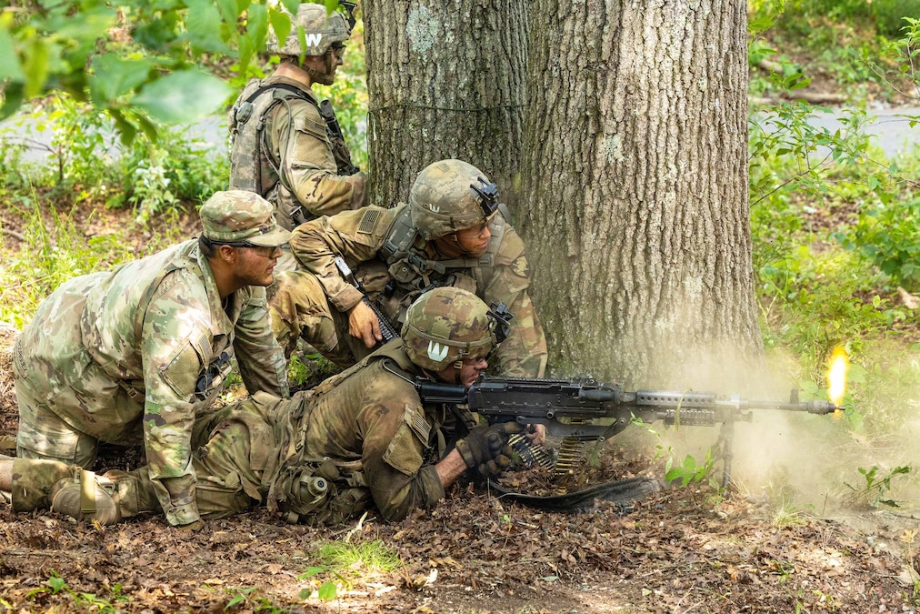 A cadet fires a weapon while lying on the ground in the forest as fellow cadets take cover behind a tree.