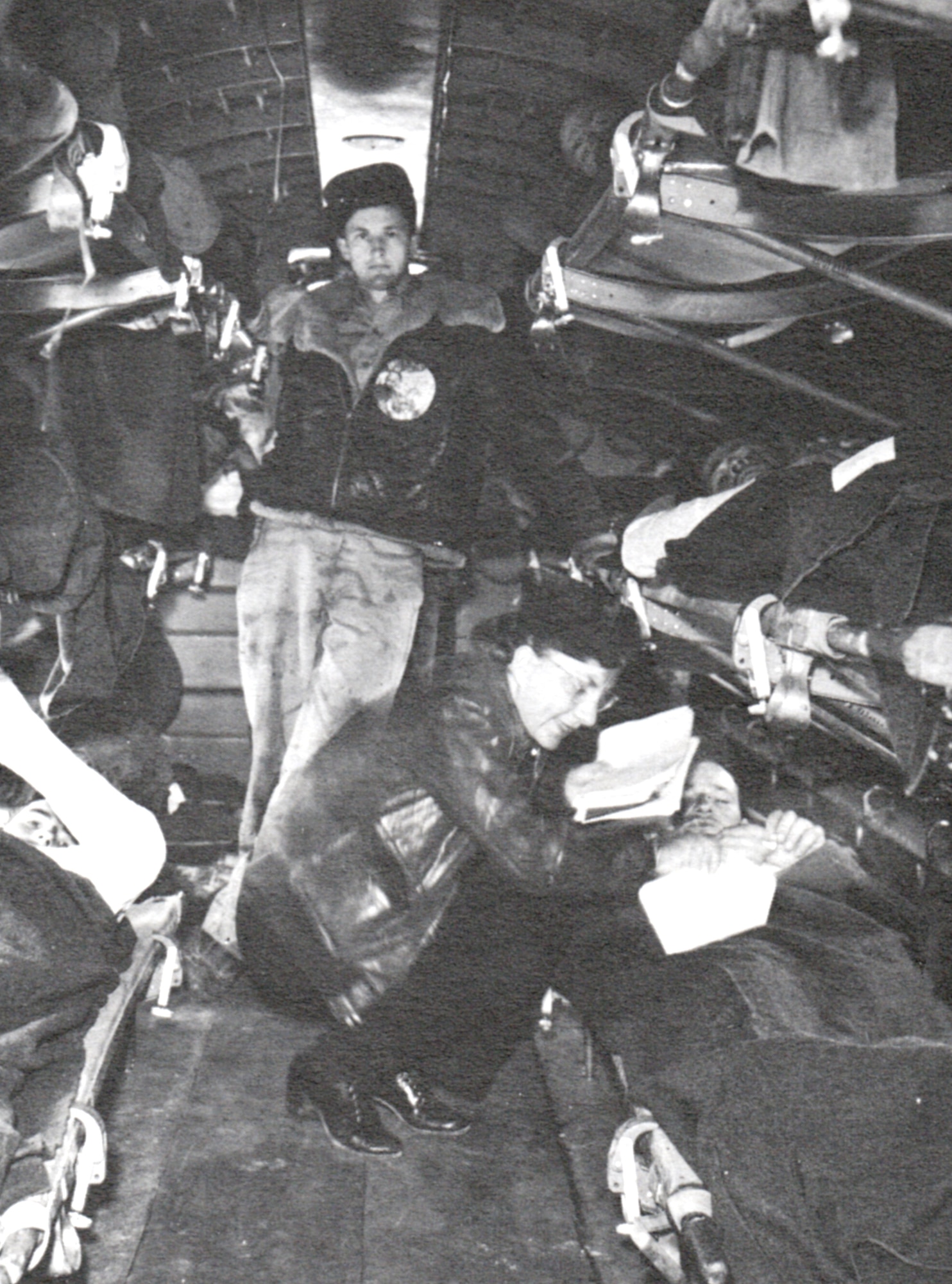 Flight nurse Lutz squatting down attending to a wounded soldier; several more soldiers are on bunks above and on the other side of the isle in the aircraft.