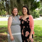 Two Caucasian women pose for a portrait photo on a sunny day in Charleston, South Carolina. The woman on the right has brown hair and is wearing a grey dress, the woman on the left has black hair and is wearing a black dress, with pink flowers on it and a pink sweater.