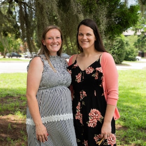 Two Caucasian women pose for a portrait photo on a sunny day in Charleston, South Carolina. The woman on the right has brown hair and is wearing a grey dress, the woman on the left has black hair and is wearing a black dress, with pink flowers on it and a pink sweater.