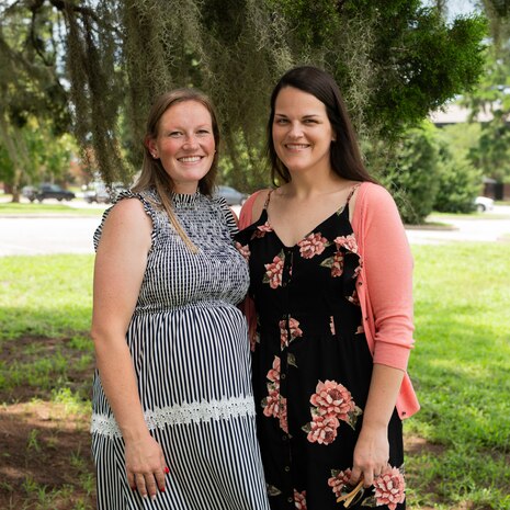 Two Caucasian women pose for a portrait photo on a sunny day in Charleston, South Carolina. The woman on the right has brown hair and is wearing a grey dress, the woman on the left has black hair and is wearing a black dress, with pink flowers on it and a pink sweater.