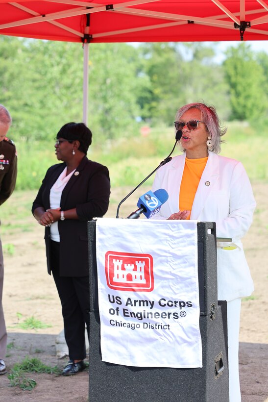 Women in white suit stands behind a podium to deliver remarks at ribbon cutting ceremony under a red canopy.