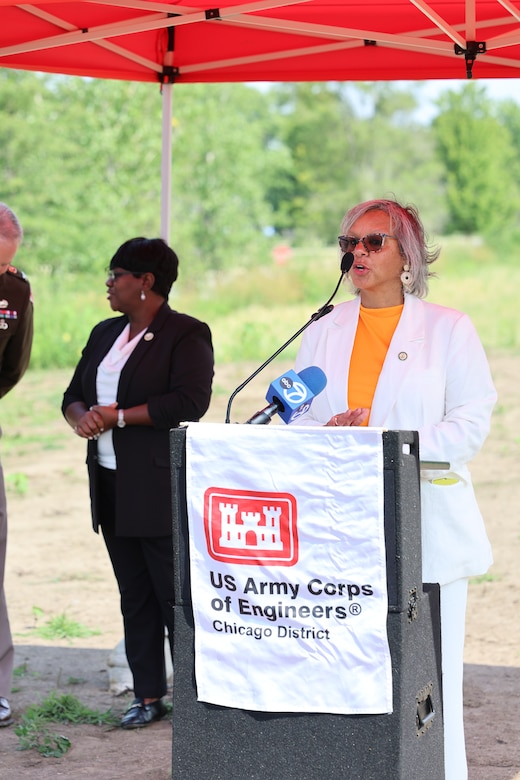 Women in white suit stands behind a podium to deliver remarks at ribbon cutting ceremony under a red canopy.