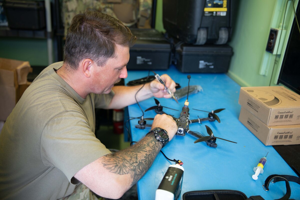 A man holds a tool and sits at a table, repairing a drone.