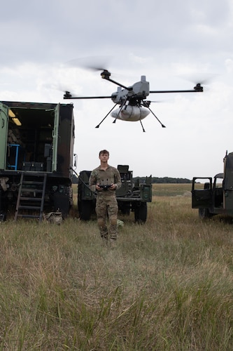 A man wearing a camouflage uniform stands in a field operating a drone. Three military vehicles are behind him.