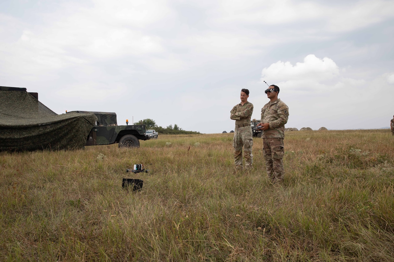 Two men wearing camouflage uniforms stand in a field, operating a drone; one of them is wearing goggles.