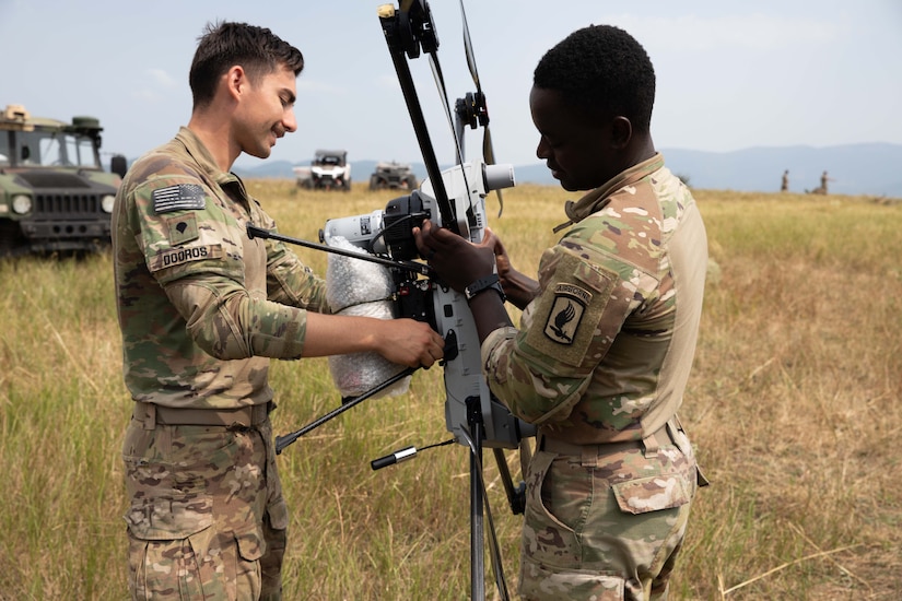Two men wearing camouflage uniforms stand in a field holding a drone.