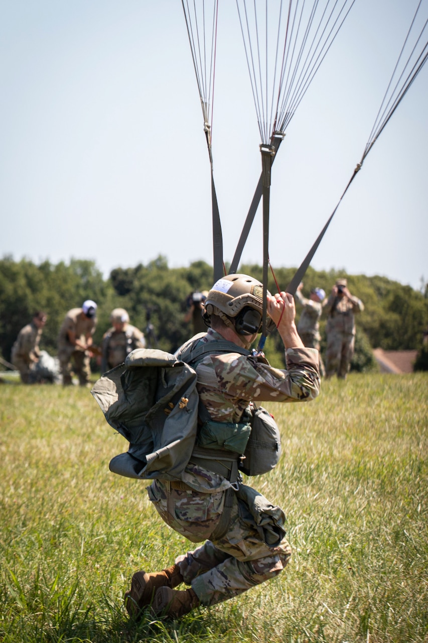 A paratrooper lands on a grassy field as others stand in the background on a sunny day.