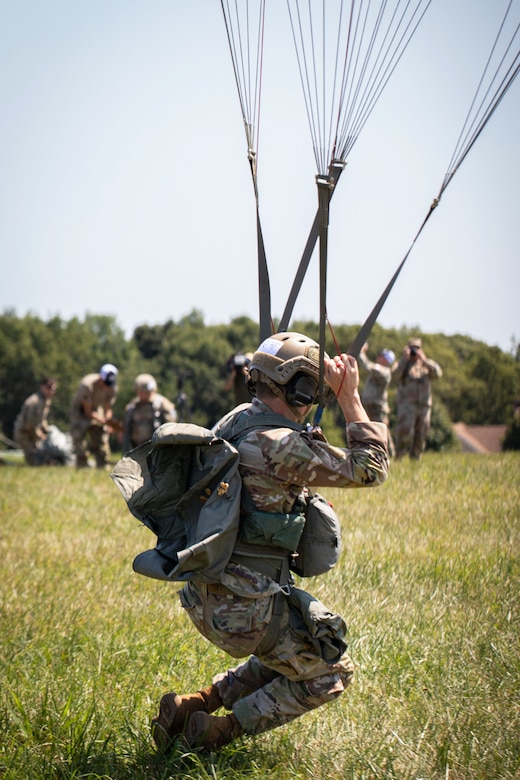 A paratrooper lands on a grassy field as others stand in the background on a sunny day.