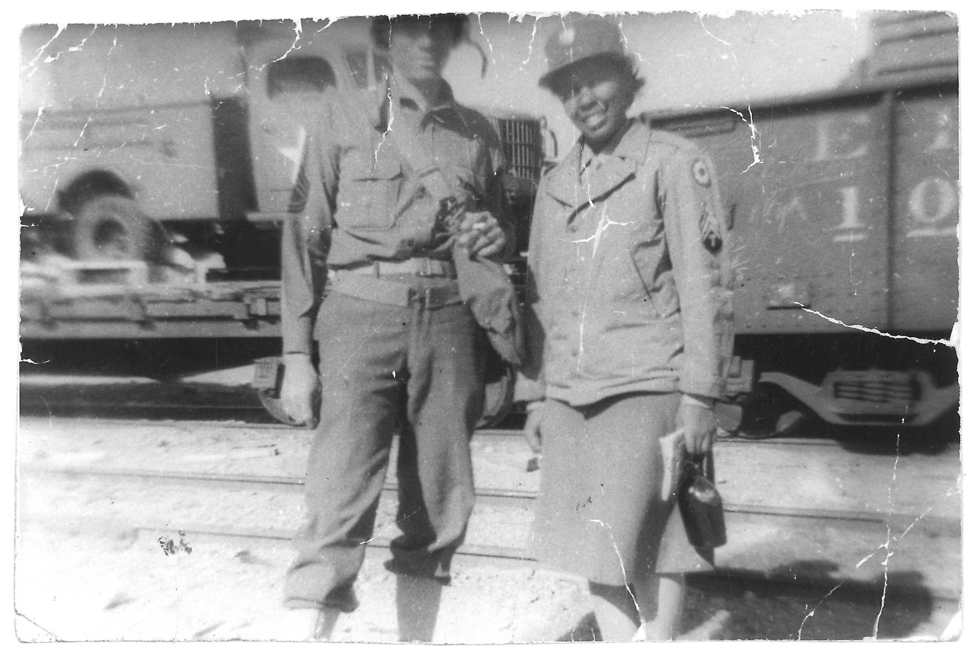 Fannie in her uniform standing next to a man in uniform; they are both standing in front of a train that is carrying a military truck