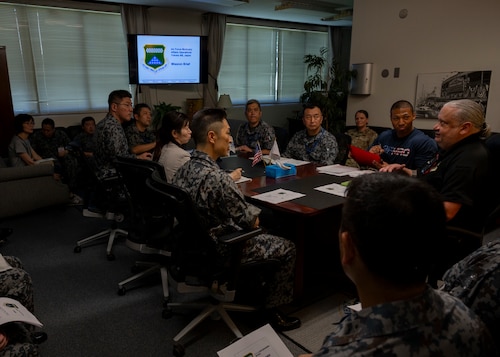 U.S. military members, civilians and Japan Air Self-Defense Force personnel listen to a brief in a conference room.