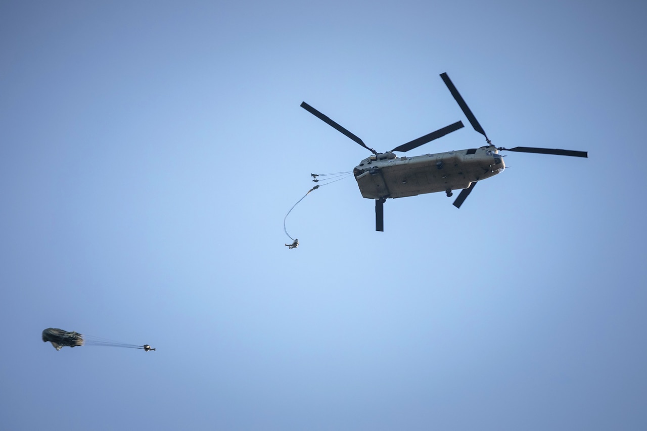 Two paratroopers are seen jumping from a helicopter, with a cloudless blue sky in the background.