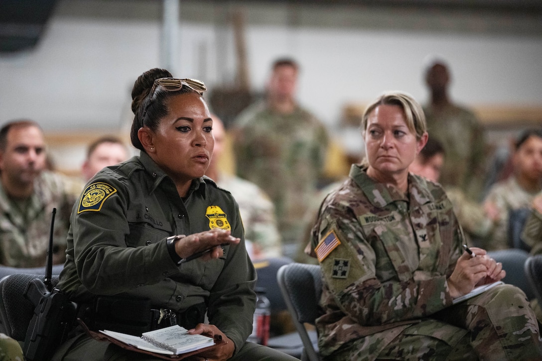 A seated Border Patrol supervisor gestures with a hand while speaking, with a soldier sitting nearby and other soldiers sitting or standing in the background.
