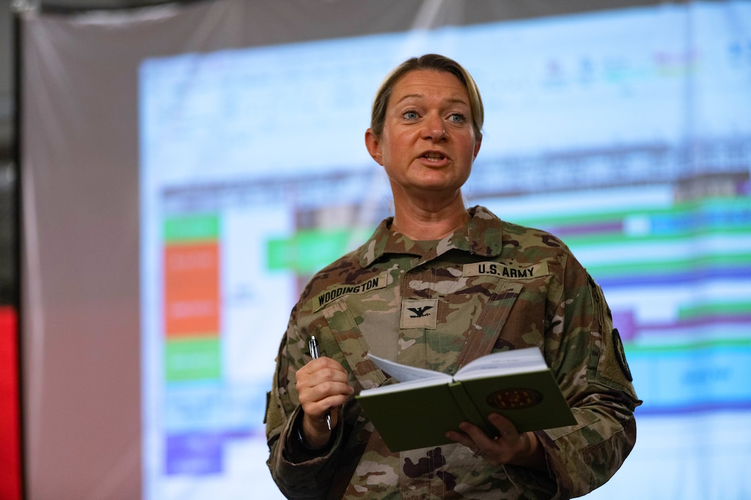 A standing soldier holds a pen and book while speaking in front of an enlarged display.