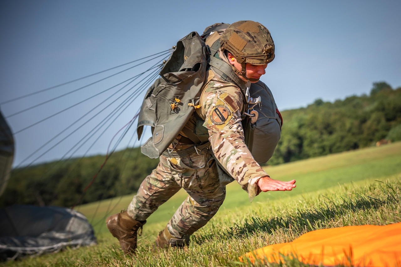 A paratrooper falls forward on a grassy field with the parachute behind the paratrooper; trees are in the background.