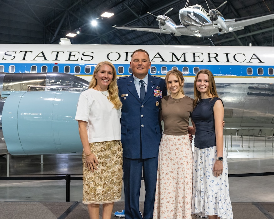A man, wearing Air Force blues service dress uniform, stands alongside three women in dresses in front of a large white plane with the text States of America visible