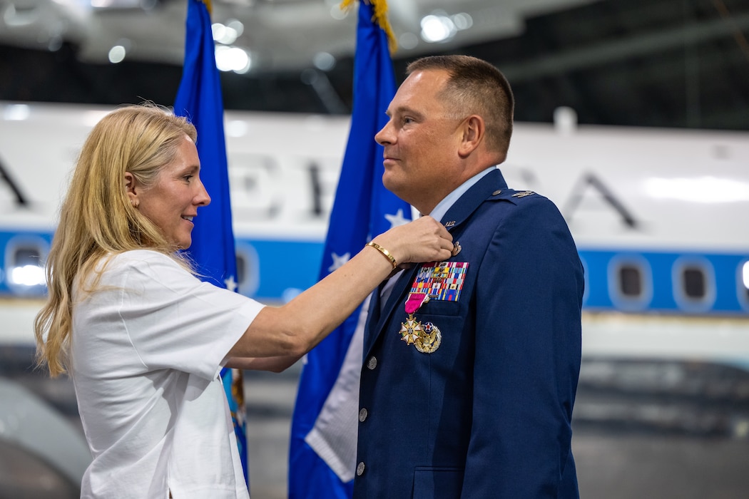 A woman, wearing a white blouse, is reaching up to place a pin on the lapel of a man, wearing Air Force blues service dress uniform