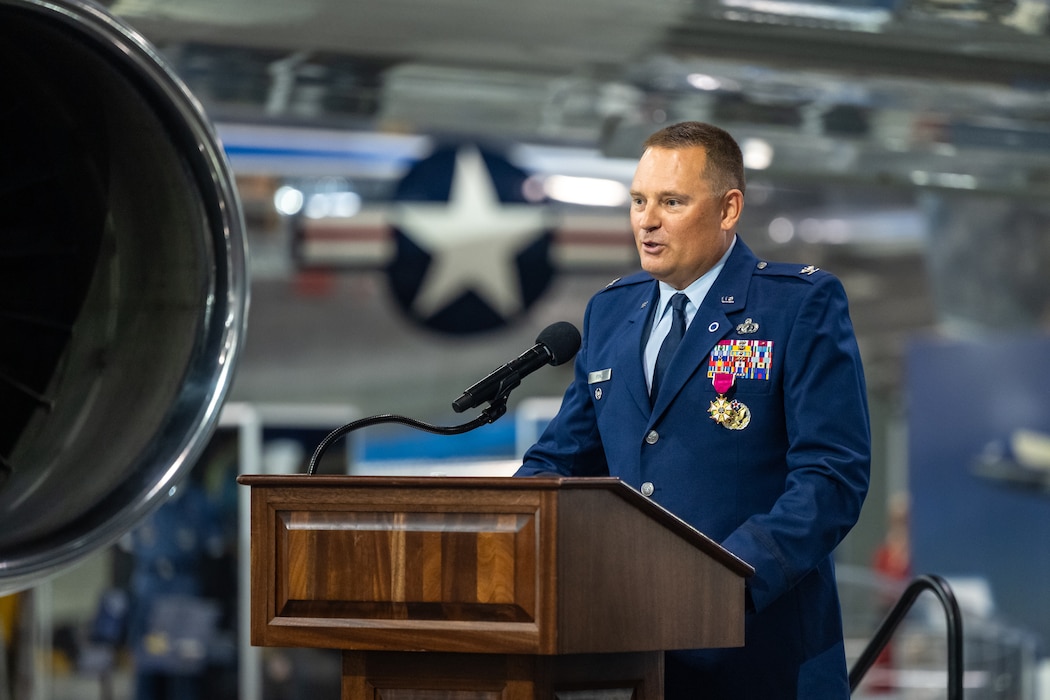 A man, wearing an Air Force blues service-dress uniform, stands behind a microphone and podium