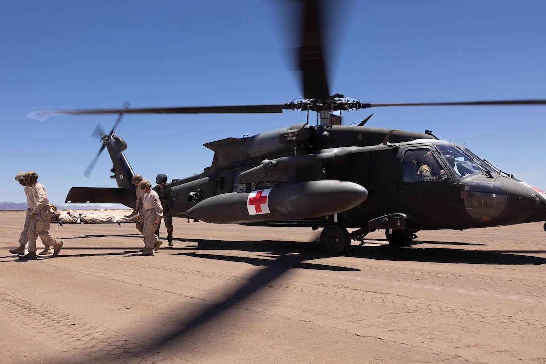 Four Marines and sailors carry a stretcher with an object on it away from a helicopter on a sandy area, as another person stands nearby on a sunny day.