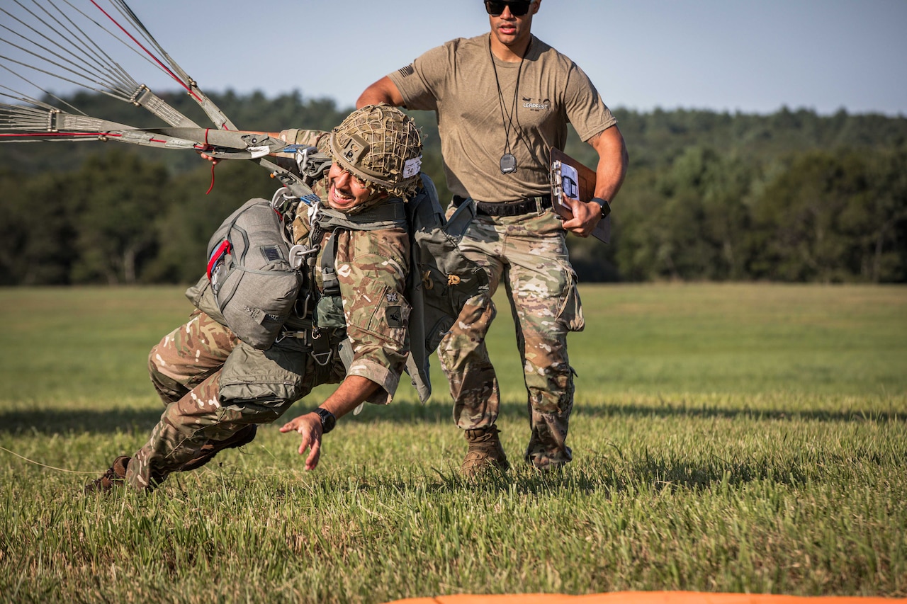 A paratrooper falls toward a pad on a grassy field, as another person holding a clipboard stands behind the paratrooper; trees are in the background.