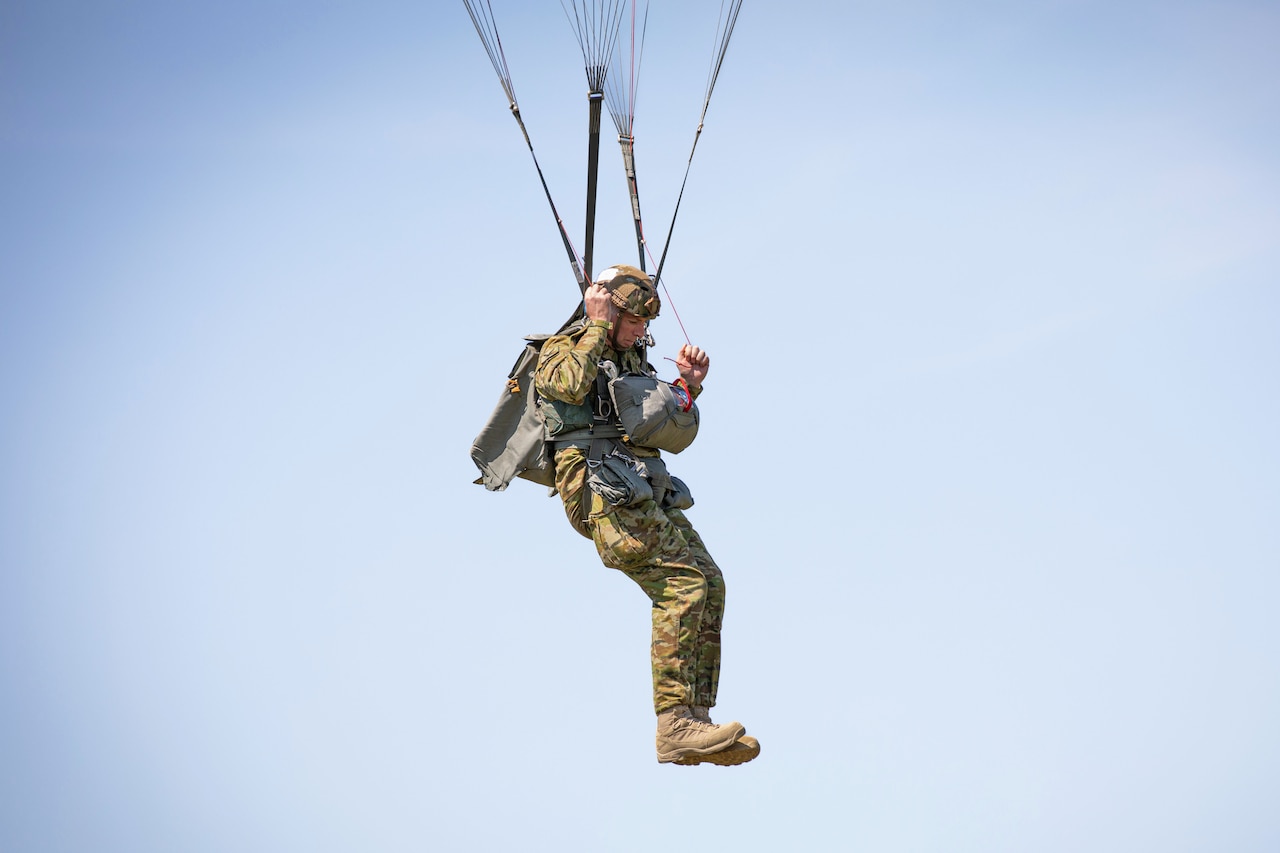 A paratrooper pulls on cords attached to a parachute and descends on a cloudless, sunny day.