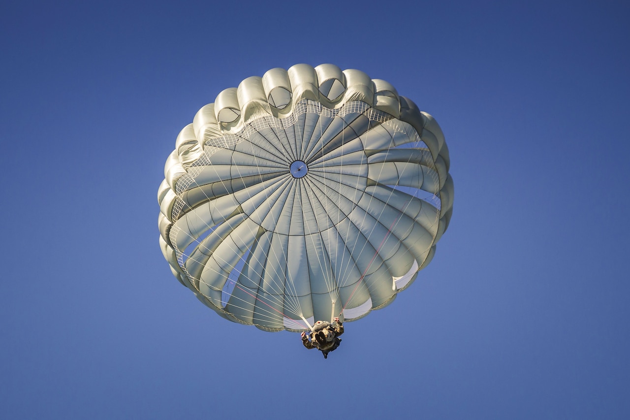 A paratrooper pulls on cords attached to a round parachute and descends on a cloudless, sunny day.