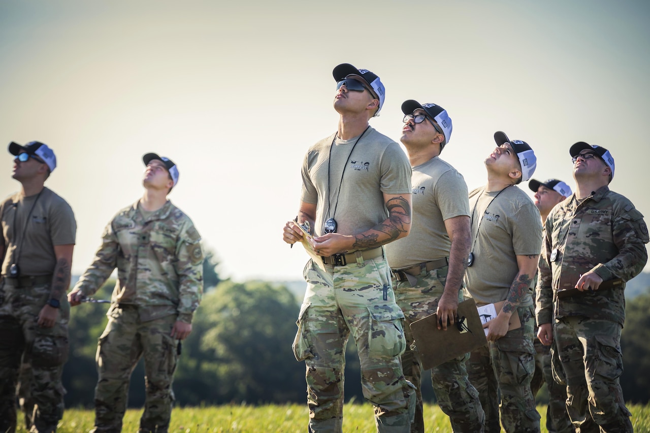 Seven military service members wearing caps stand in a grassy field and look skyward on a sunny day.
