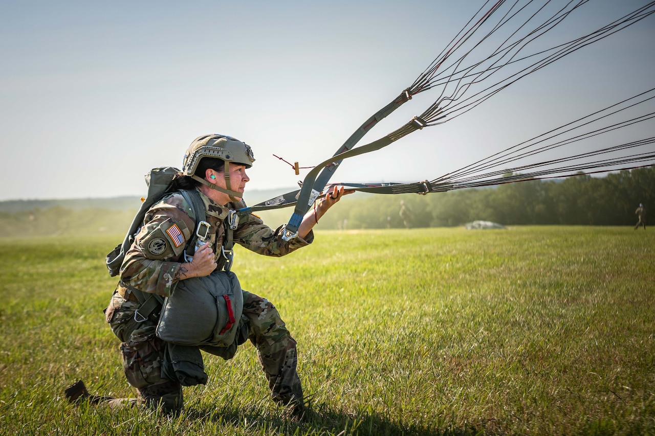 A soldier kneels on a grassy field and pulls at a strap of an attached parachute blowing in the opposite direction of the soldier.