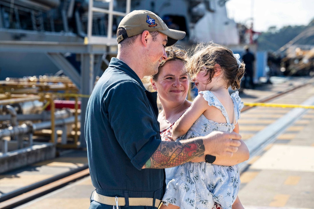 A sailor, wife and daughter hug on a pier during the day.