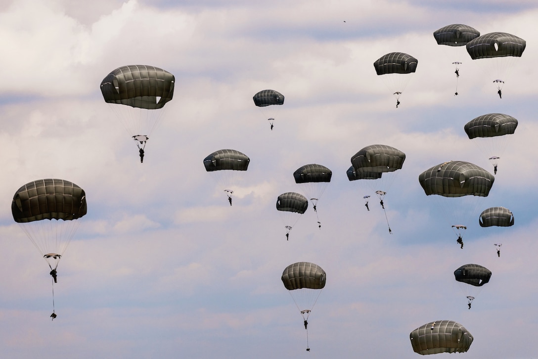 More than a dozen soldiers with parachutes descend against a cloudy, blue sky.