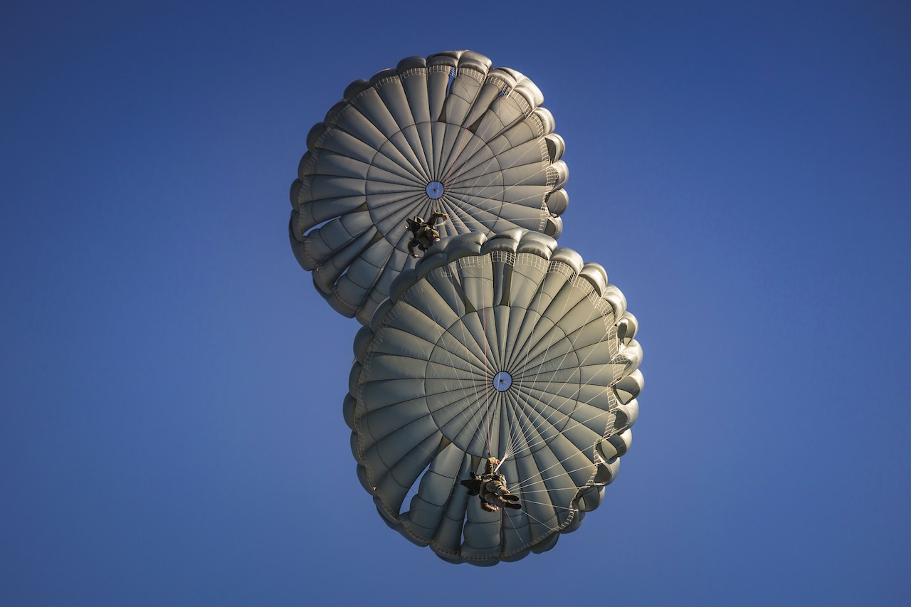 A pair of paratroopers descend near each other in a blue sky, with their parachutes billowing above them.