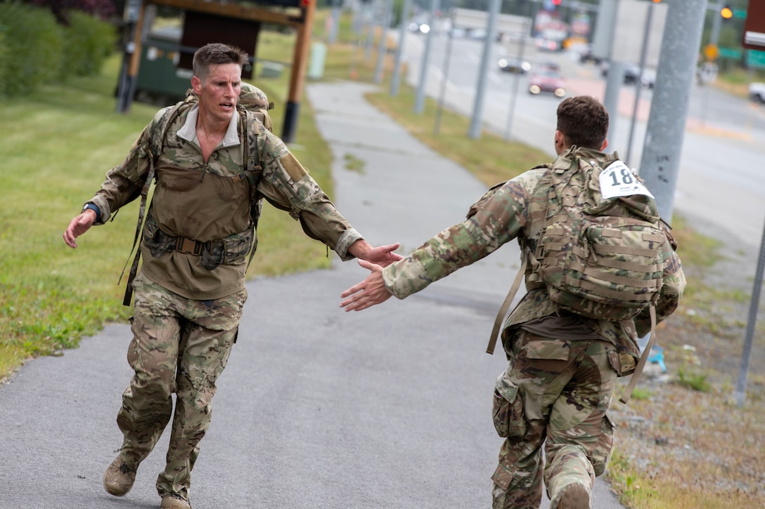 Service members wearing backpacks slap hands while running past each other near a highway.