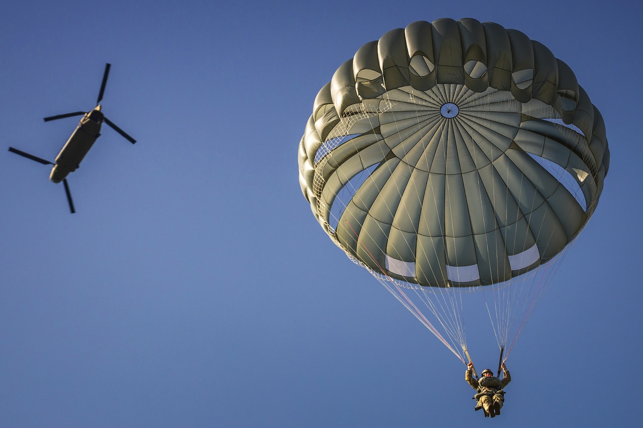 A paratrooper descends in a blue sky, with the parachute billowing above the paratrooper and a helicopter flying overhead.