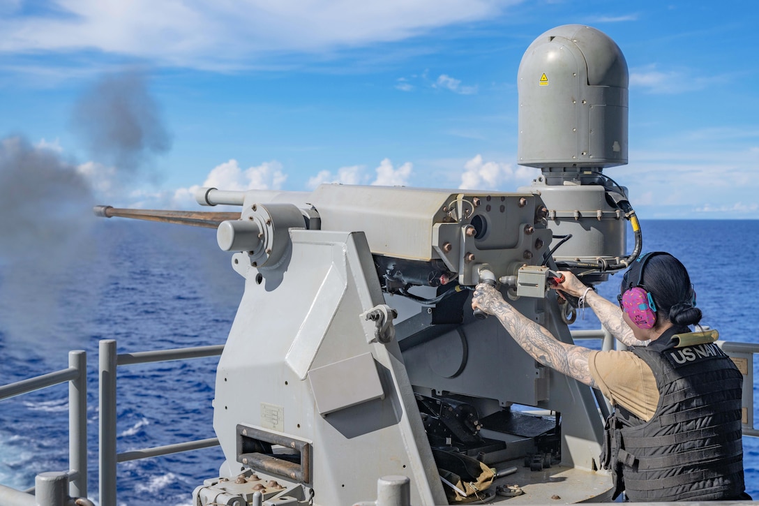 A sailor wearing a bullet proof vest fires a weapon aboard a ship at sea creating clouds of smoke.