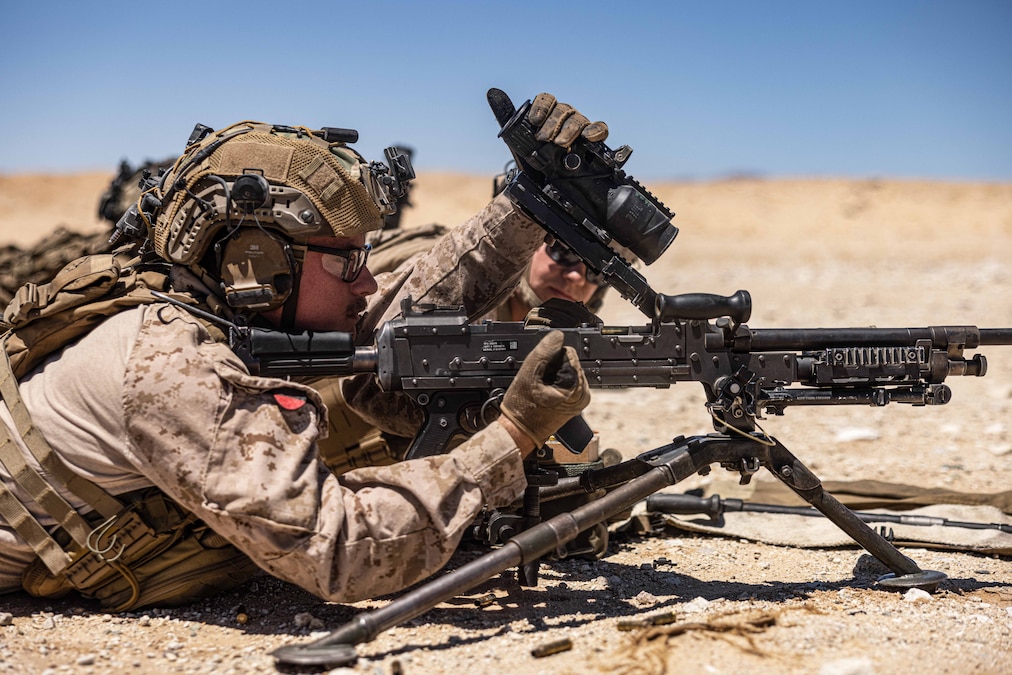 A Marine works on a weapon while laying on the ground in a desert-like area.