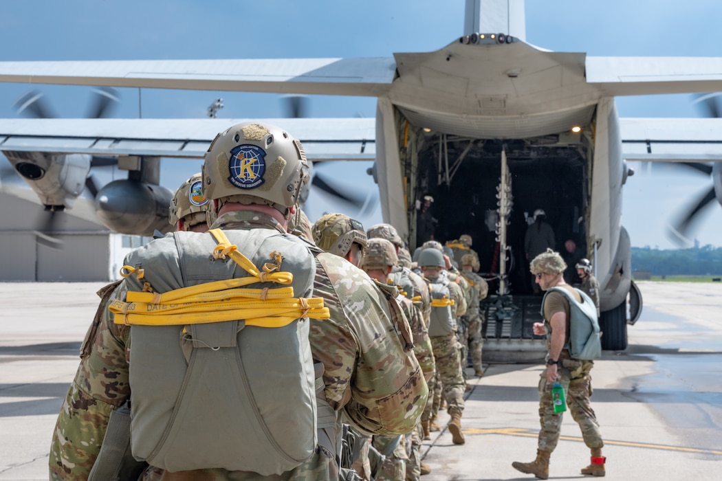 A shot from behind a single file line of Army soldiers wearing camouflage uniforms, helmets, and parachute packs leading up to the open bay of a military aircraft