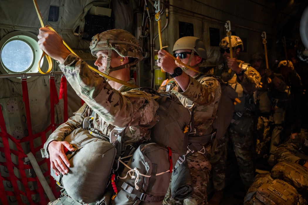A line of Army soldiers wearing helmets and parachute packs hold onto yellow ropes attached from above