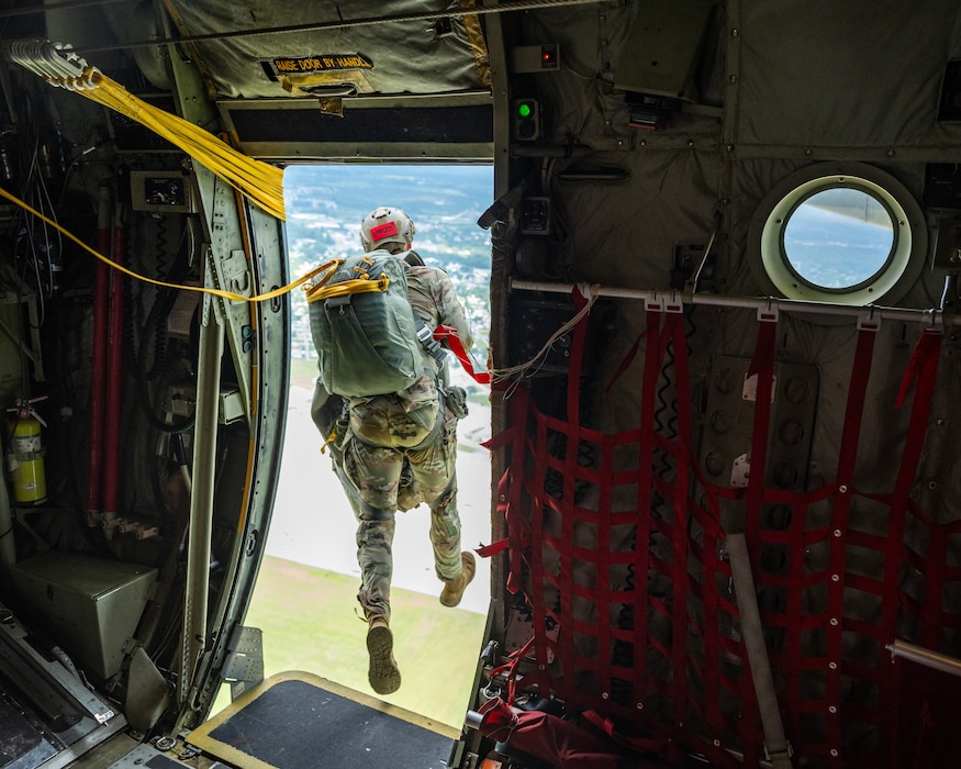 A person, wearing Army camouflage uniform and large parachute pack, steps out of an open plane door. Green and grey ground can be seen in the background