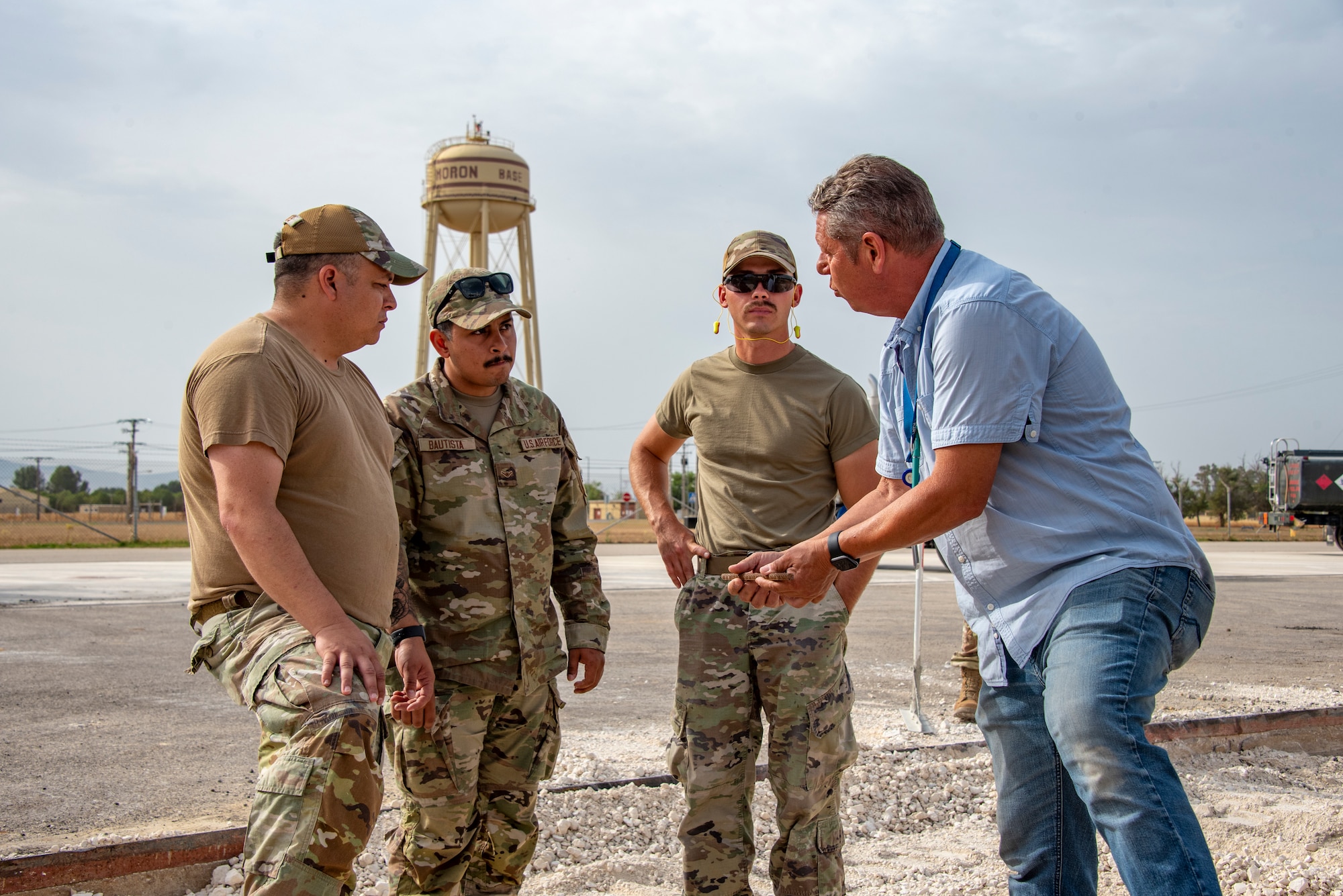 Three uniformed Airmen speak with a Spanish contractor at a construction site with the Moron Air Base water tower in the distance.