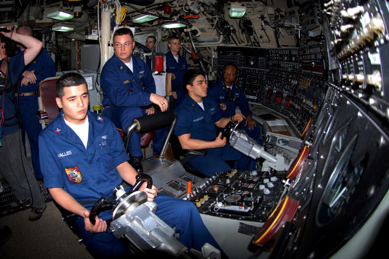 Eight people wearing blue military uniforms operate controls inside a submarine.
