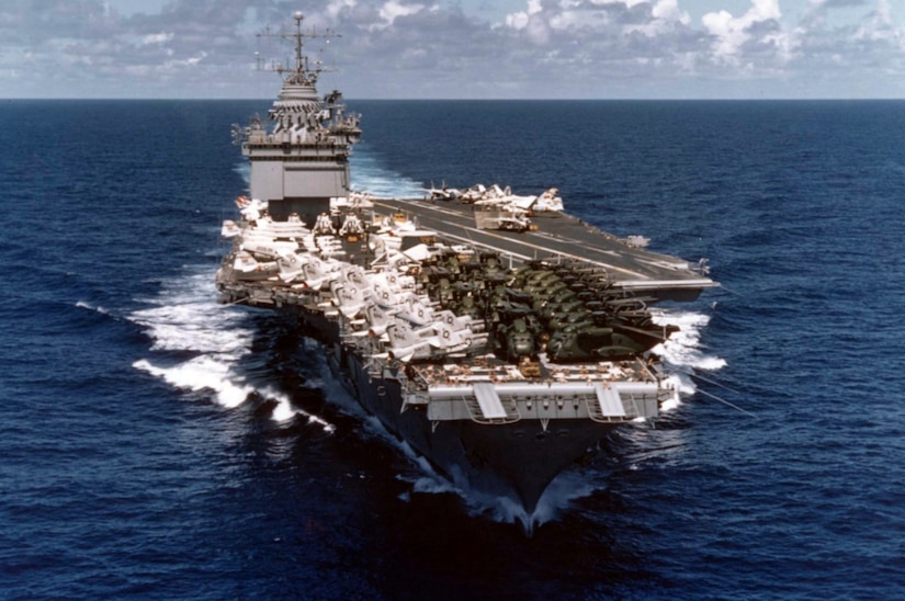 An aircraft carrier with airplanes on its deck moves through the ocean.