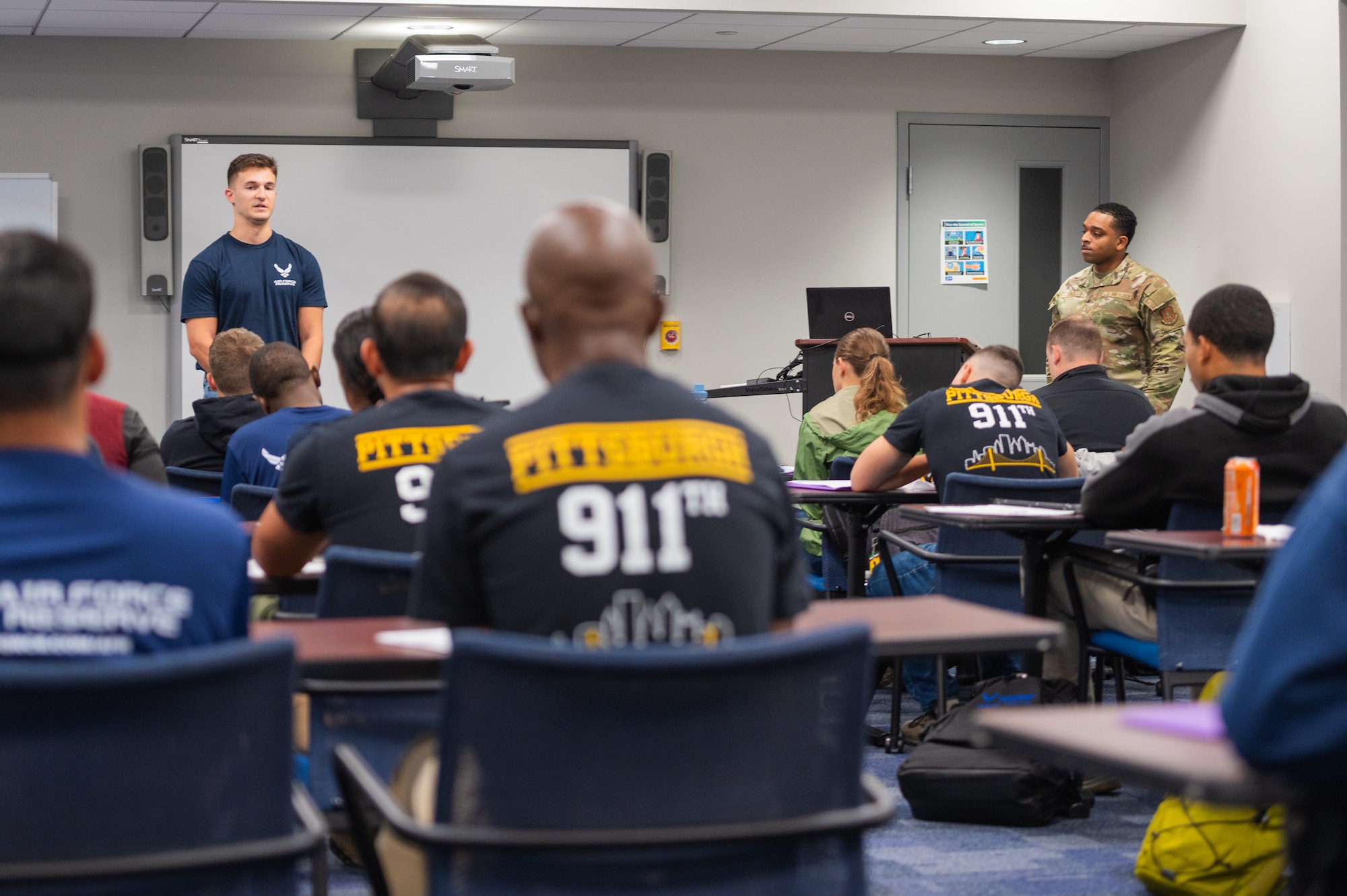 a white male in a blue air force t-shirt speaks in front of a classroom filled with recruits