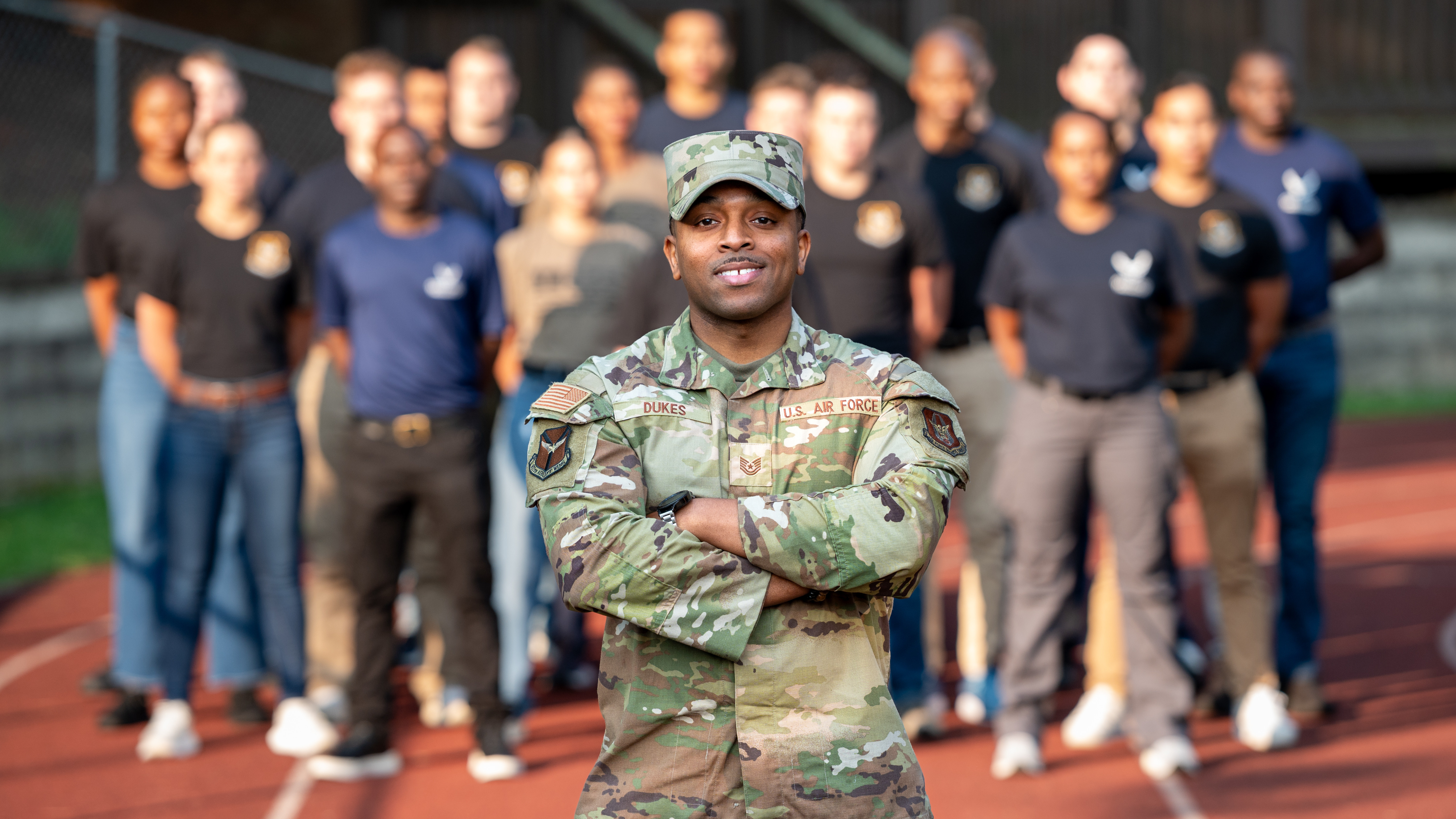 an African American male in a camo uniform stands with his arms crossed in front of a group of new recruits.
