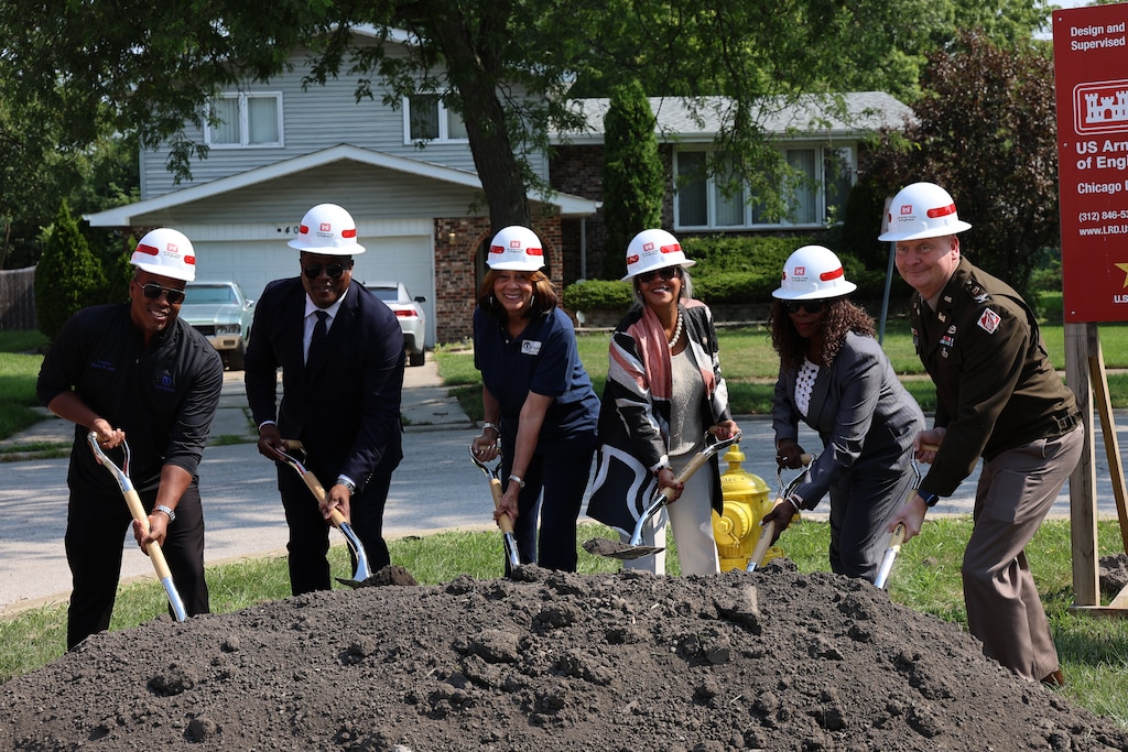 Six people stand over a mound of dirt all wearing white hardhats and using a shovel during groundbreaking ceremony.