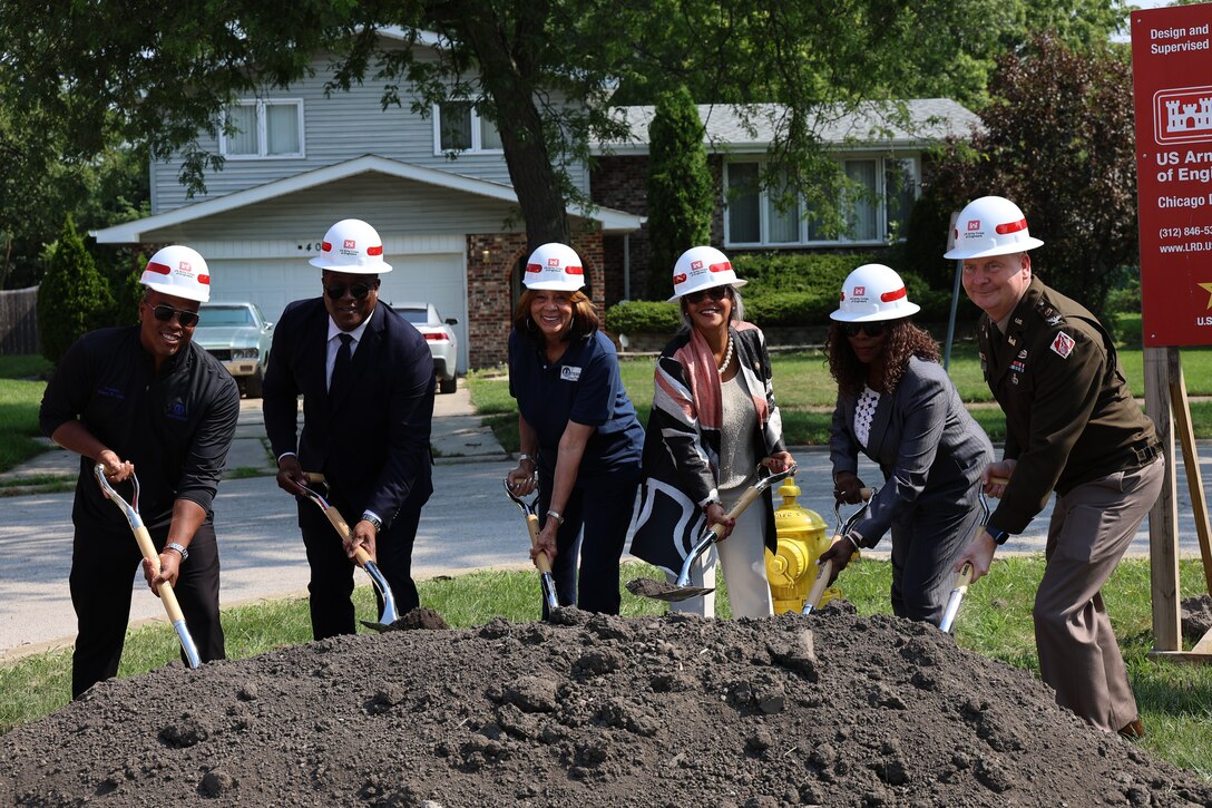 Six people stand over a mound of dirt all wearing white hardhats and using a shovel during groundbreaking ceremony.