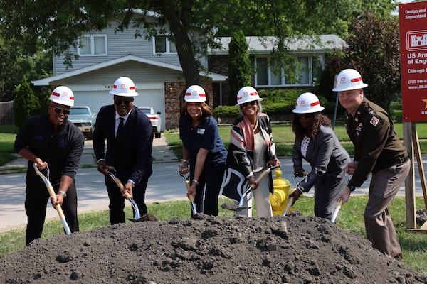 Six people stand over a mound of dirt all wearing white hardhats and using a shovel during groundbreaking ceremony.