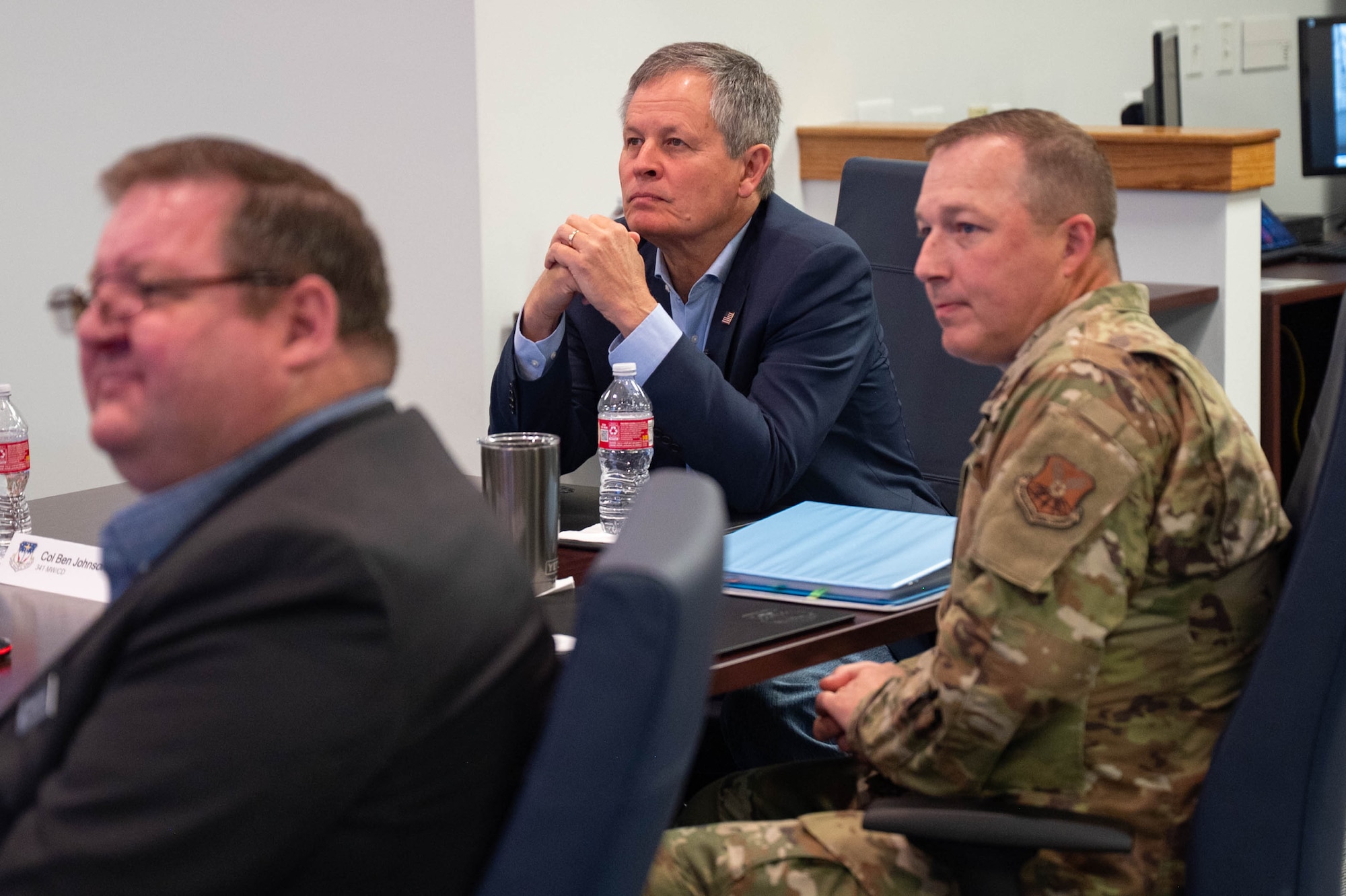 A man in civilian attire and a man in uniform sit at a table listening to a mission brief.