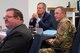 A man in civilian attire and a man in uniform sit at a table listening to a mission brief.